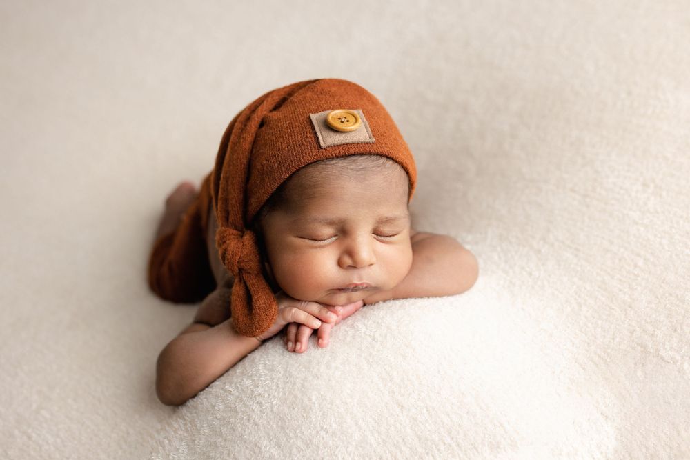 Sleeping newborn rests peacefully on white blanket wearing brown knit hat with decorative tail.