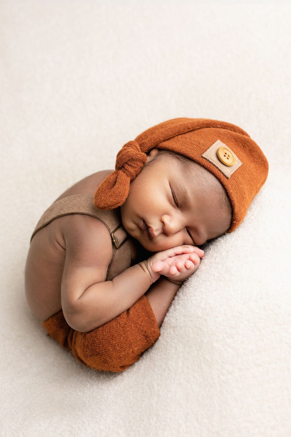 Sleeping newborn in a cozy rust-colored knotted hat and romper lying on a soft white surface.