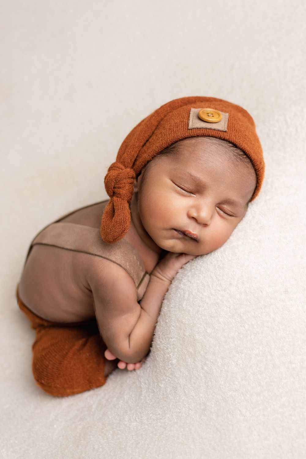 Sleeping newborn in brown knit cap and bodysuit lying peacefully on soft white fabric.