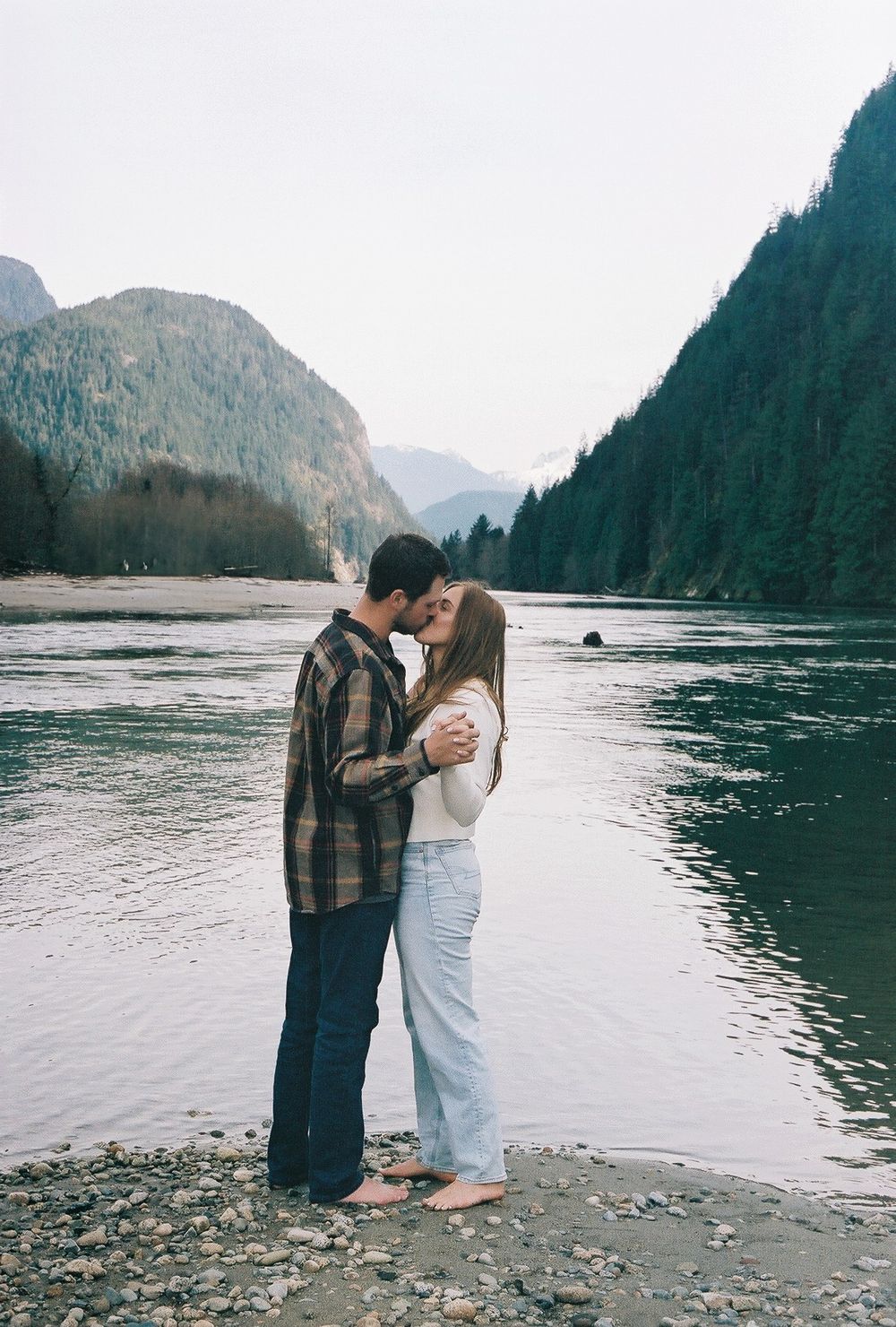 Couple sharing intimate moment on rocky beach with mountain lake views.