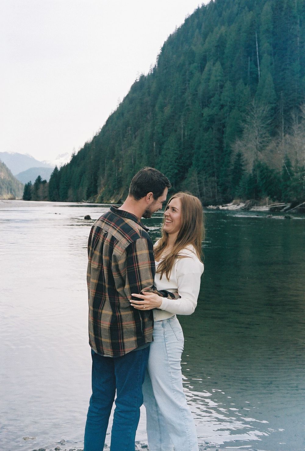 A couple shares an intimate moment by a scenic mountain lake with evergreen forests in the background.