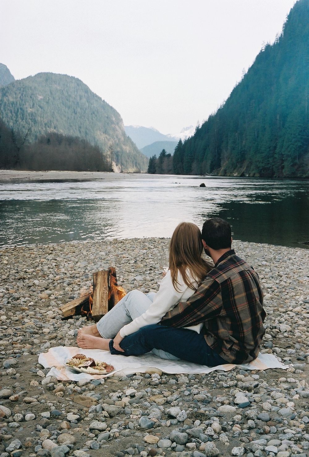 A couple relaxes on a blanket by a rocky lakeshore surrounded by mountain scenery.