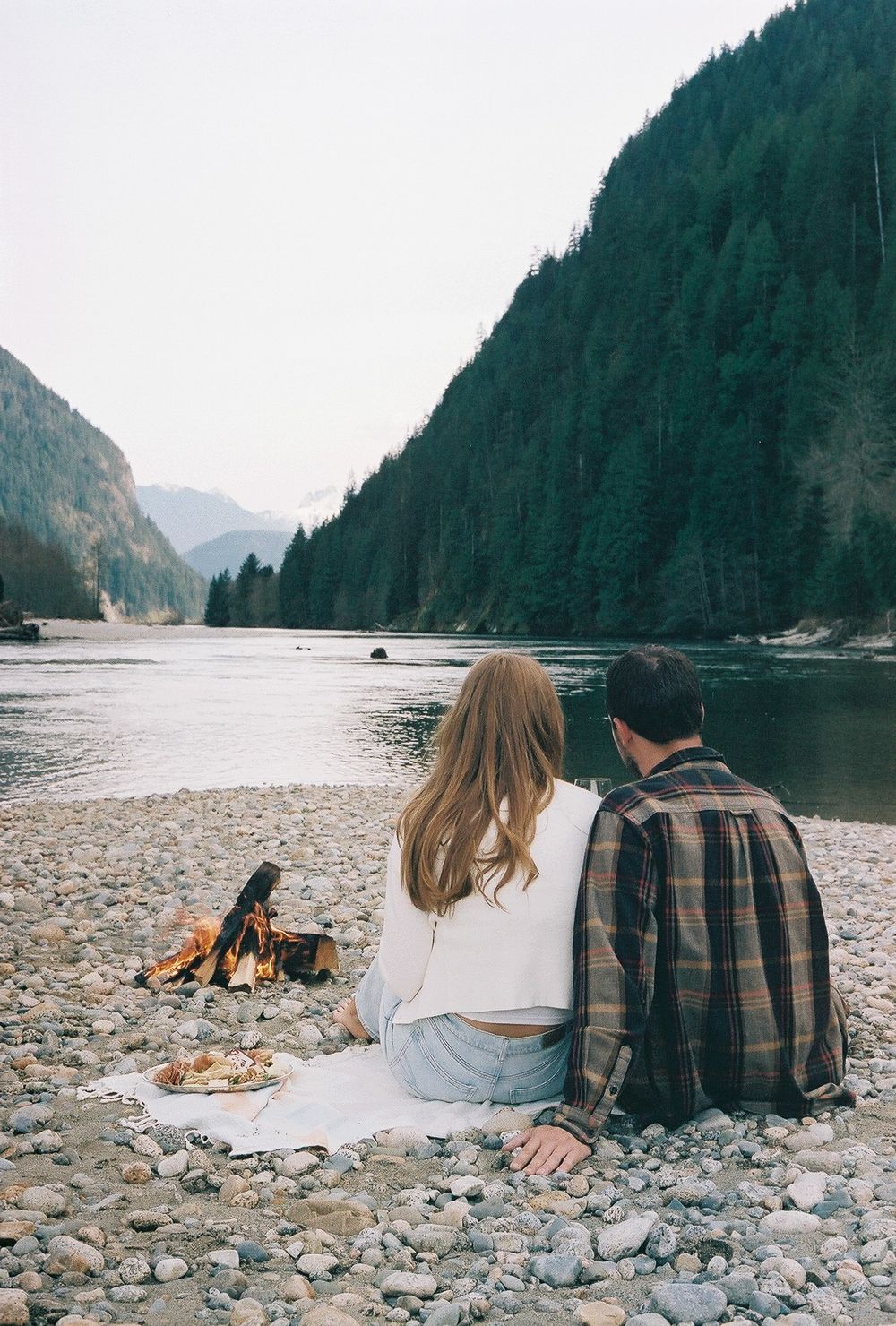 Couple sitting by campfire on rocky beach with mountains in background.