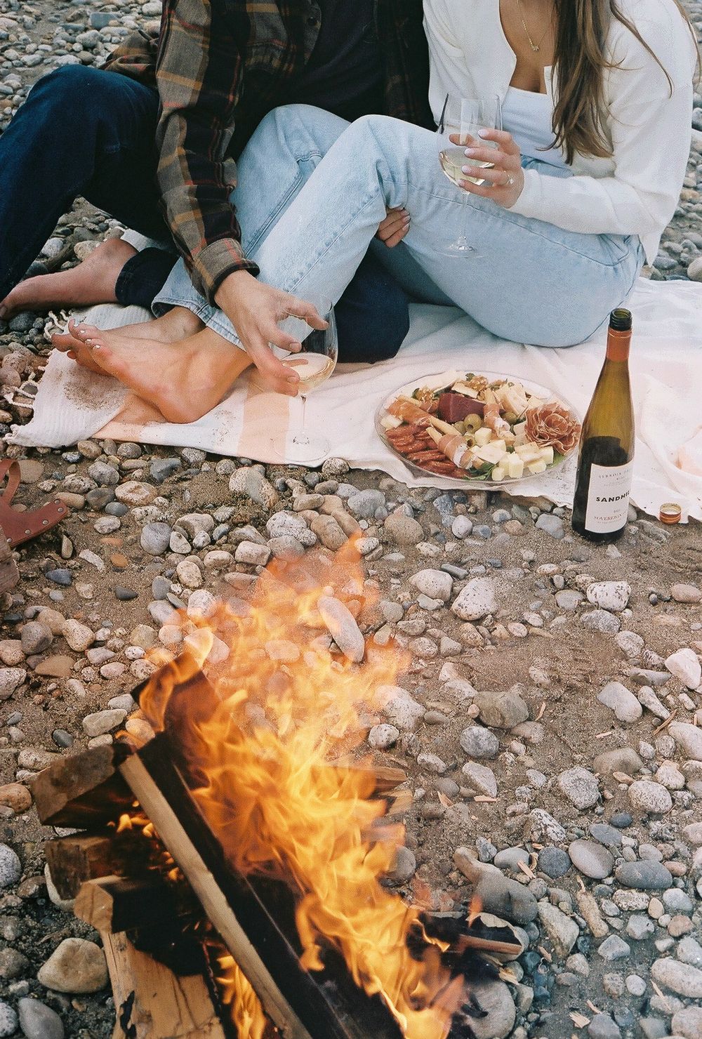 Campfire burning bright with wine and blanket setup on rocky beach.