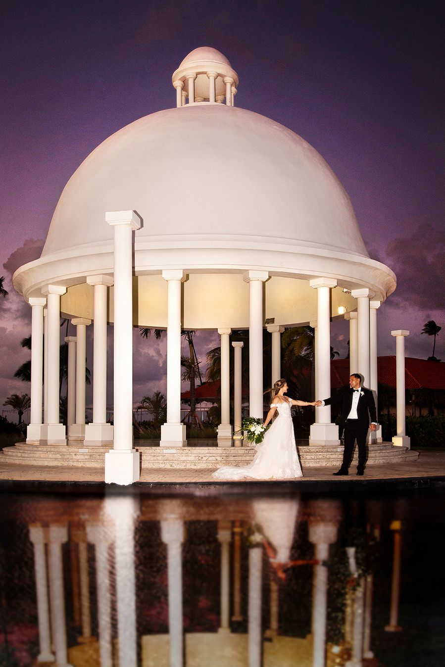 A white domed gazebo with columns reflects in a pool of water at sunset during a wedding ceremony. (Puerto Rico)