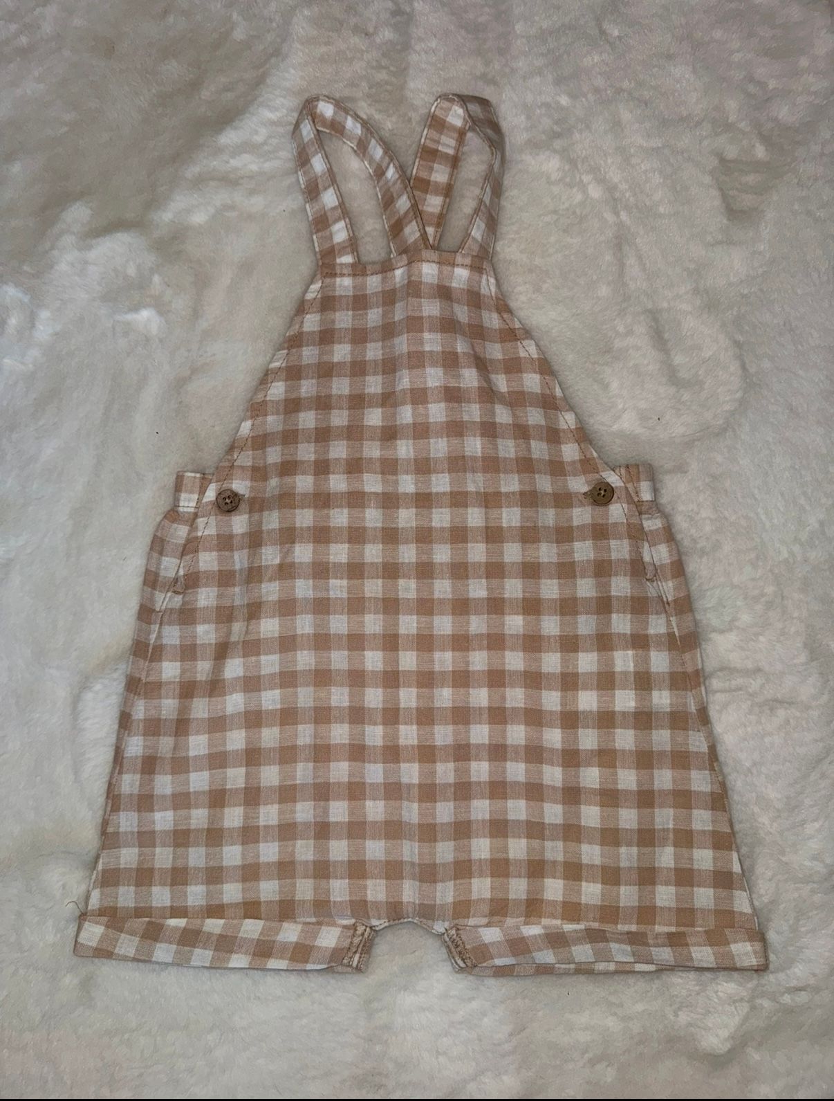 Brown and white gingham pattern baby overalls laid flat on white surface.