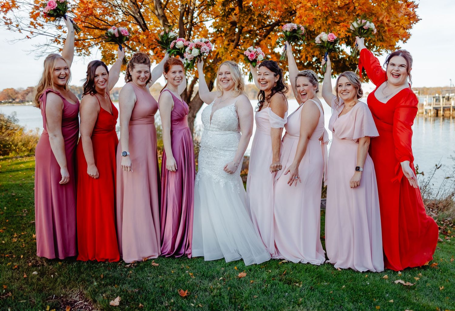 Wedding party in autumn wearing an array of pink and red dresses poses by a lake with colorful fall foliage in background.