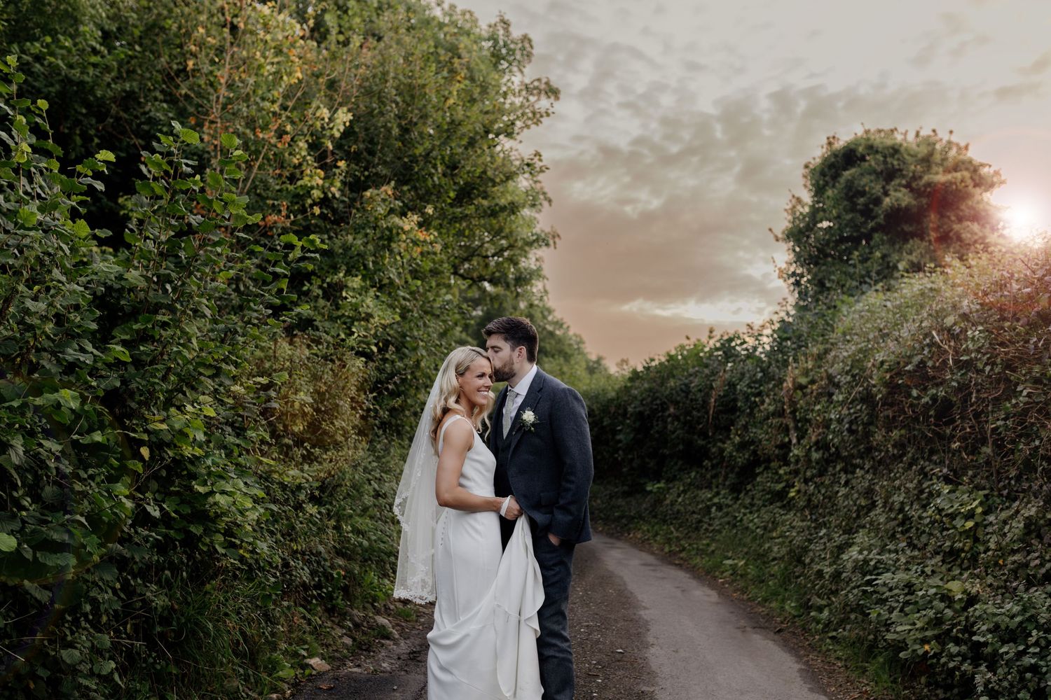 A newlywed couple embraces on a countryside path at sunset with lush greenery surrounding them.
