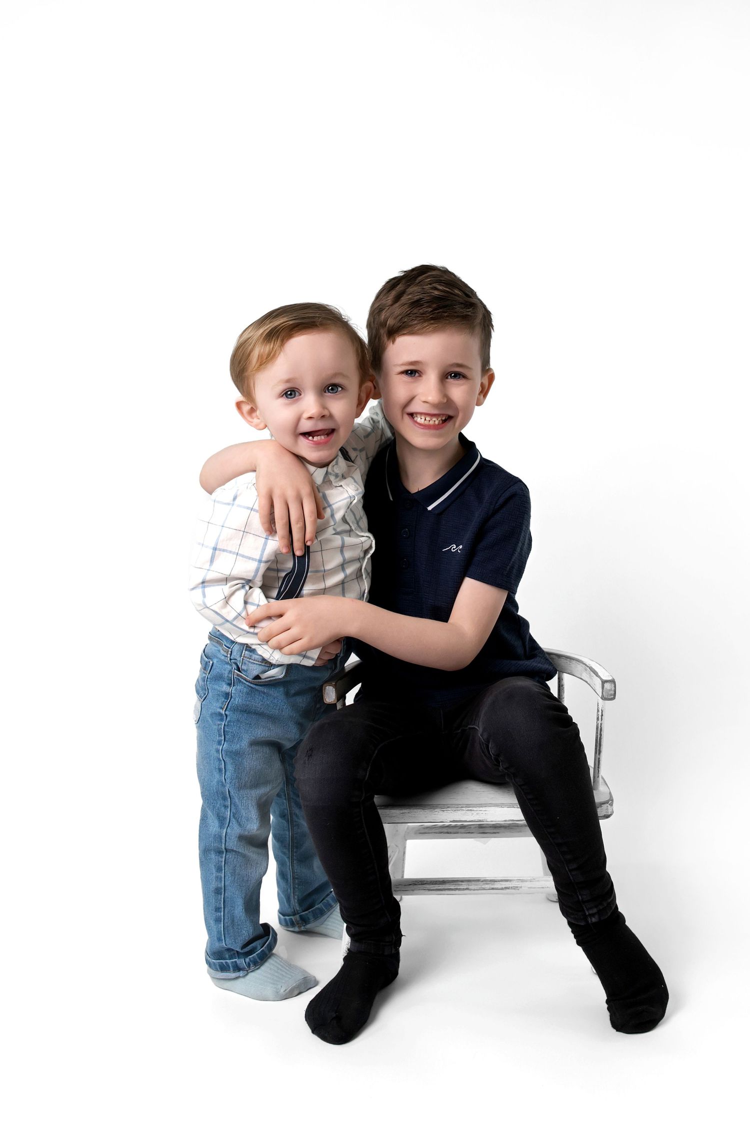 Two young children pose together on a white bench, sharing a happy moment against a plain white background.