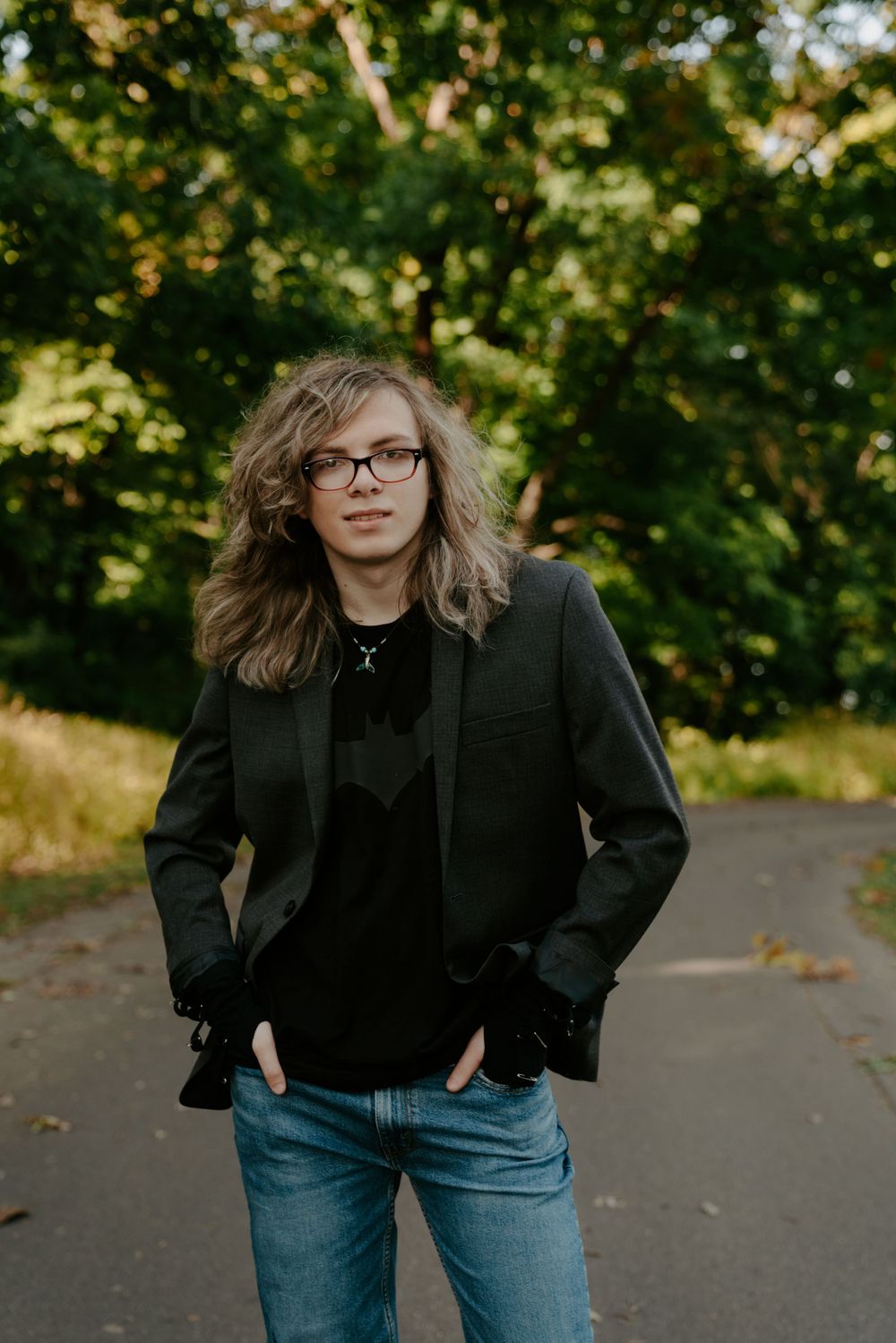Someone in a black jacket and jeans poses playfully on a tree-lined path.