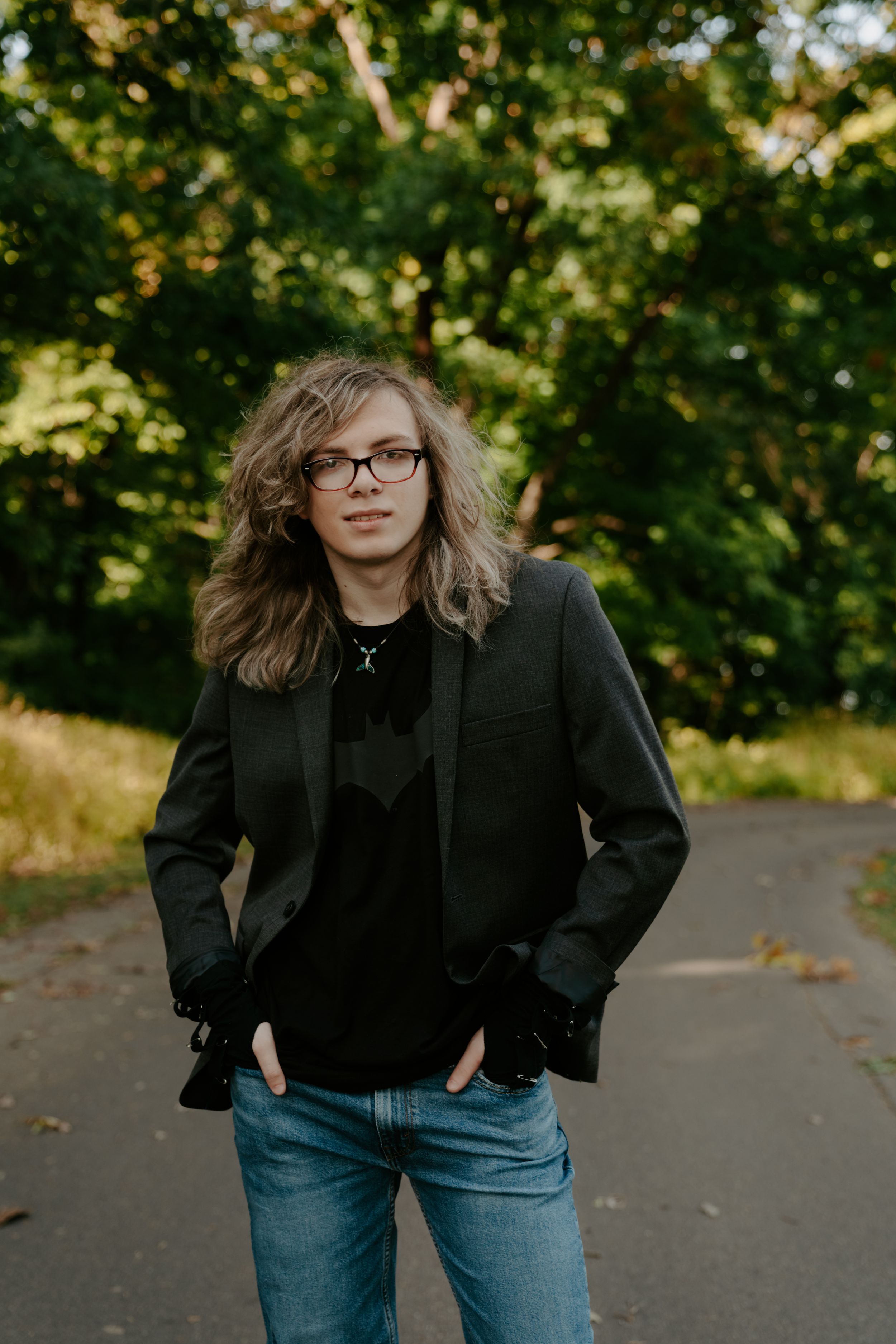 Someone in a black jacket and jeans poses playfully on a tree-lined path.
