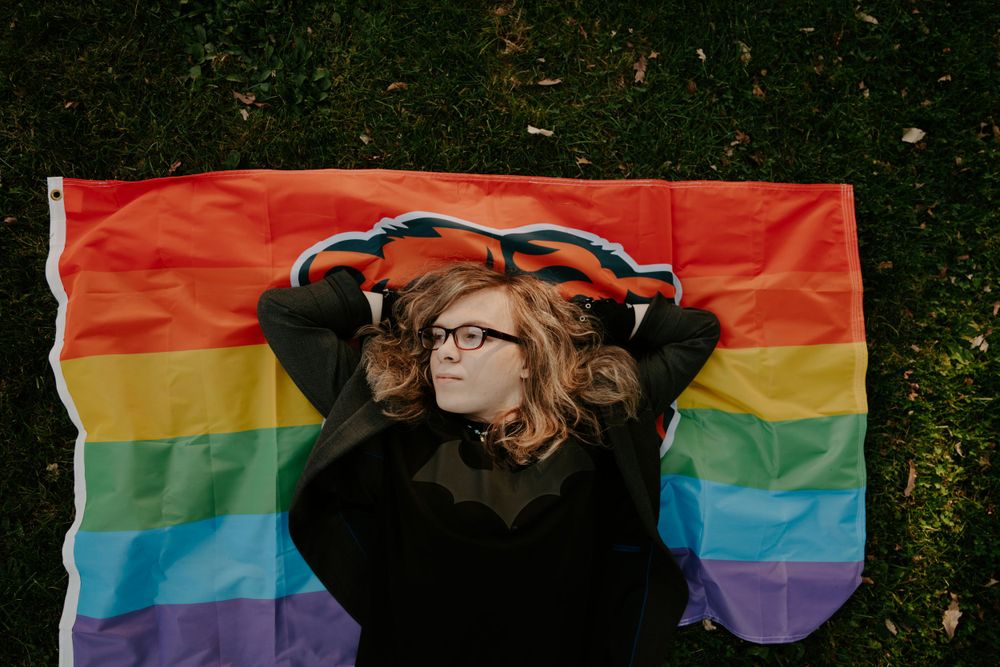 A person stands in front of a vibrant rainbow pride flag outdoors.