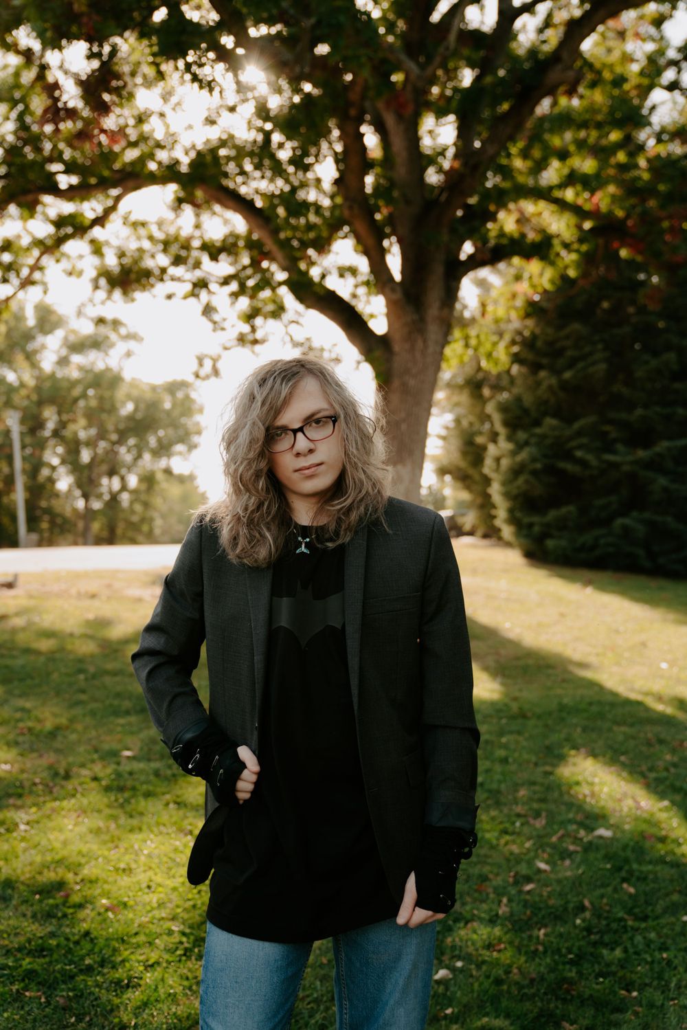 Someone in black clothing poses casually on a grassy hill with trees in the background.