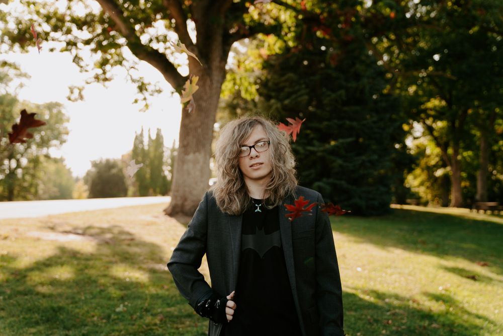 A person in dark clothing poses on a sunlit grassy slope with trees.