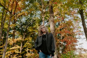 Person in black jacket standing among autumn trees in a park setting.