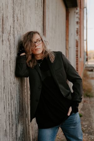 Person in black outfit posing against weathered wooden wall panels.