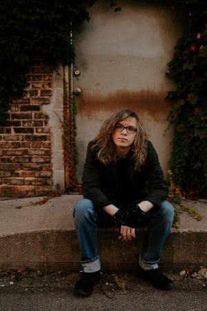 Person in black clothing sitting against brick wall in moody lighting.