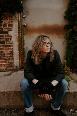 Person in casual attire sitting on ground against brick wall with ivy.