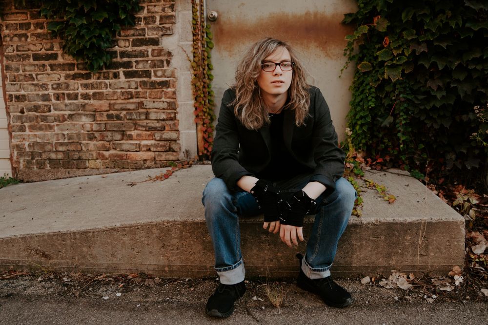 Person in dark clothing sitting on concrete against brick wall with greenery.