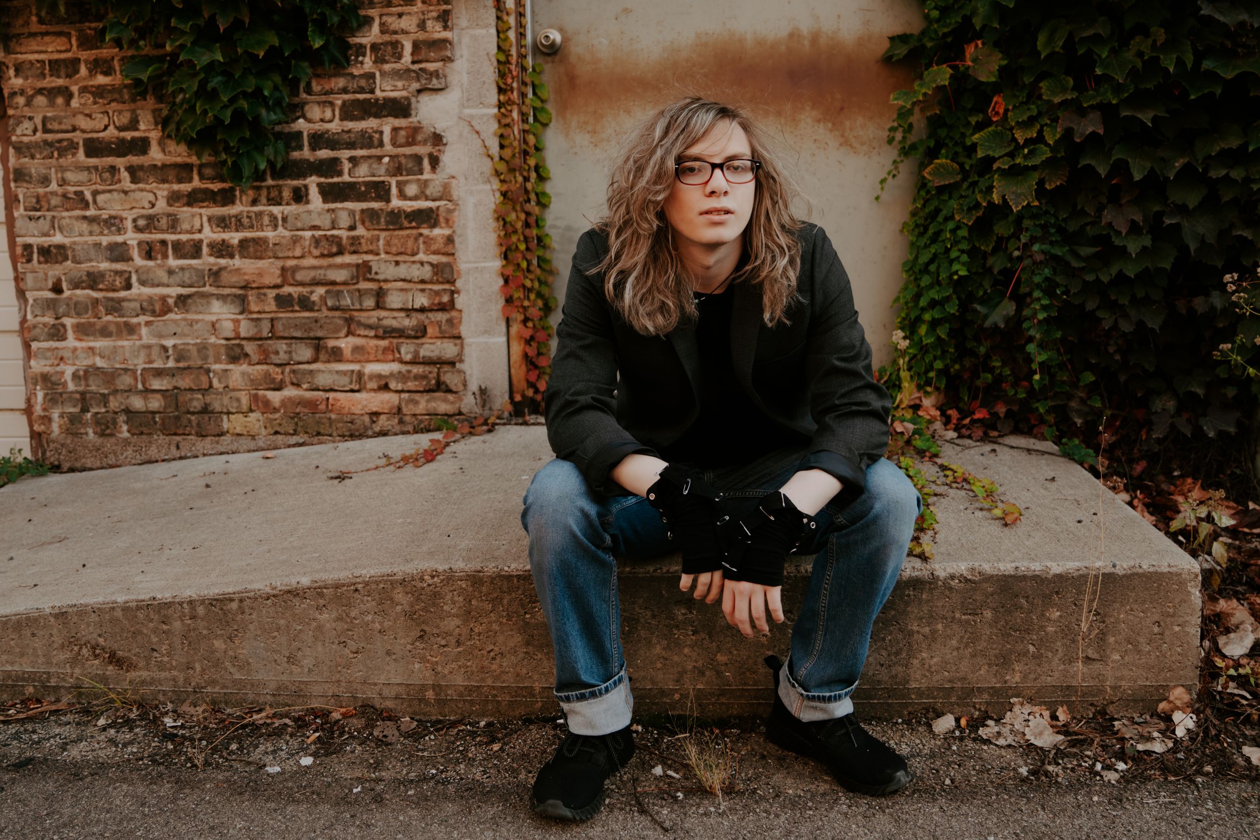 Person in dark clothing sitting on concrete against brick wall with greenery.