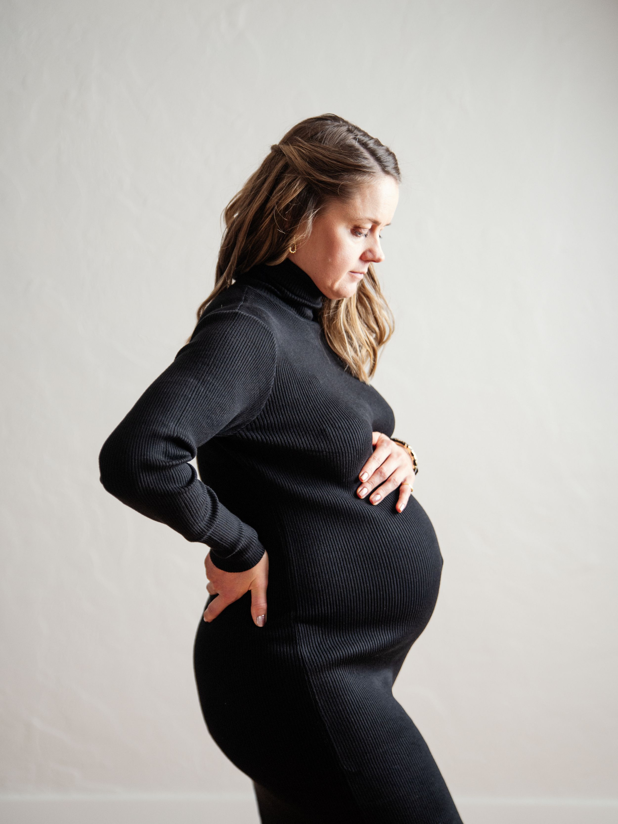 Side profile of a pregnant woman in a black ribbed sweater dress holding her baby bump against a light background.