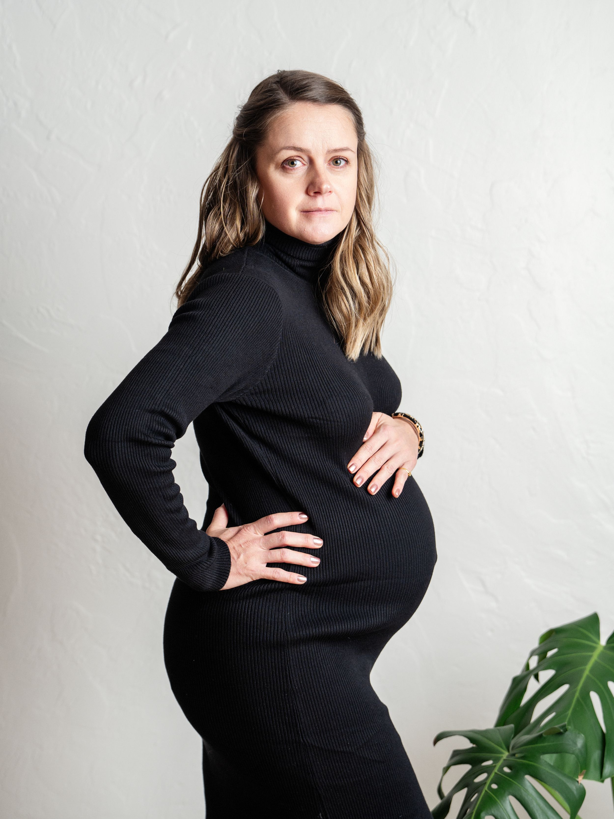 Woman in black turtleneck dress poses showing off her pregnancy bump against a white wall.