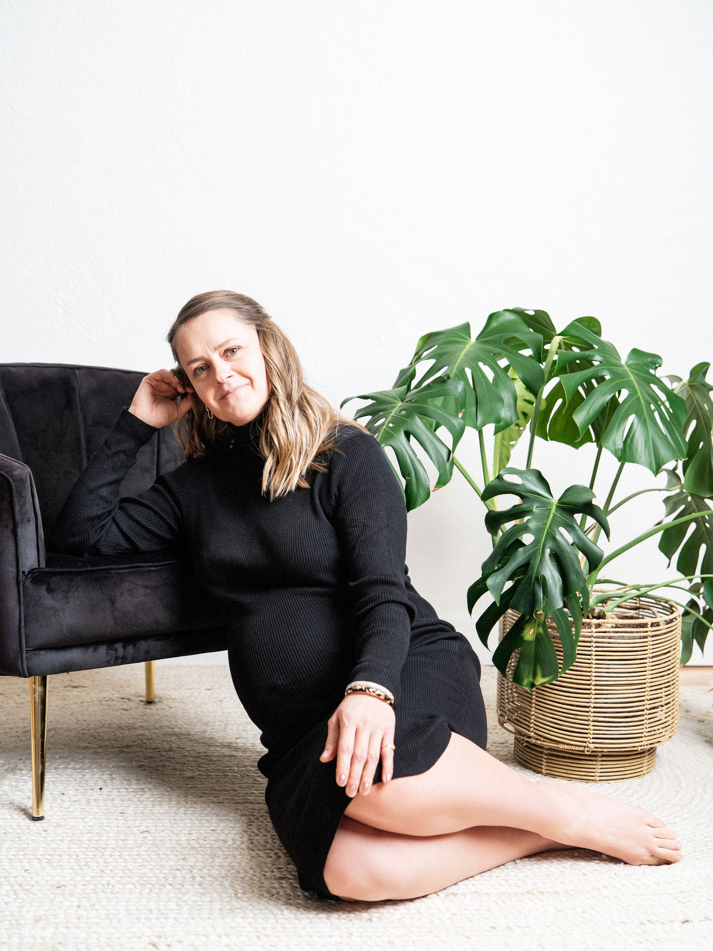 Lifestyle portrait in a minimalist interior with a black armchair, monstera plant, and relaxed figure in dark attire.