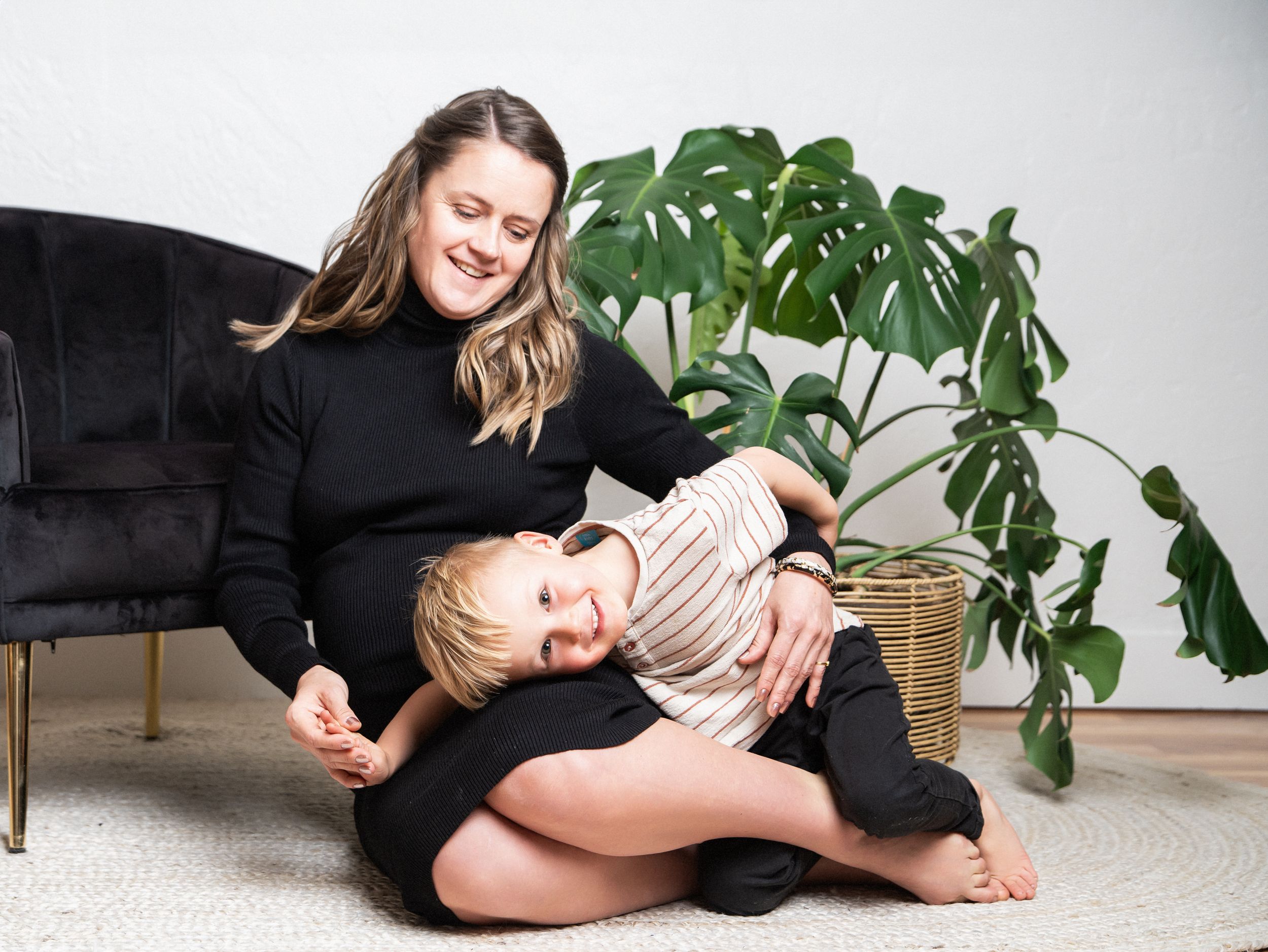 Two people sit on a grey carpet in front of a large monstera plant and black velvet couch, sharing a joyful moment together.