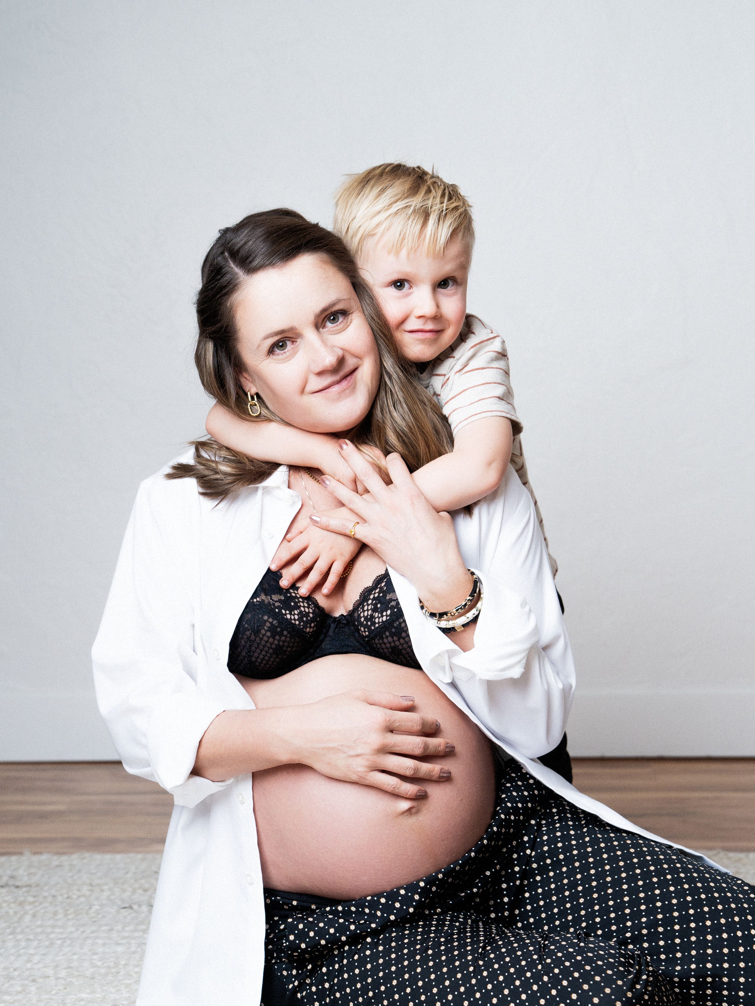 A pregnant woman in a white shirt and polka dot pants embraces a young child on a carpeted floor against a gray backdrop.
