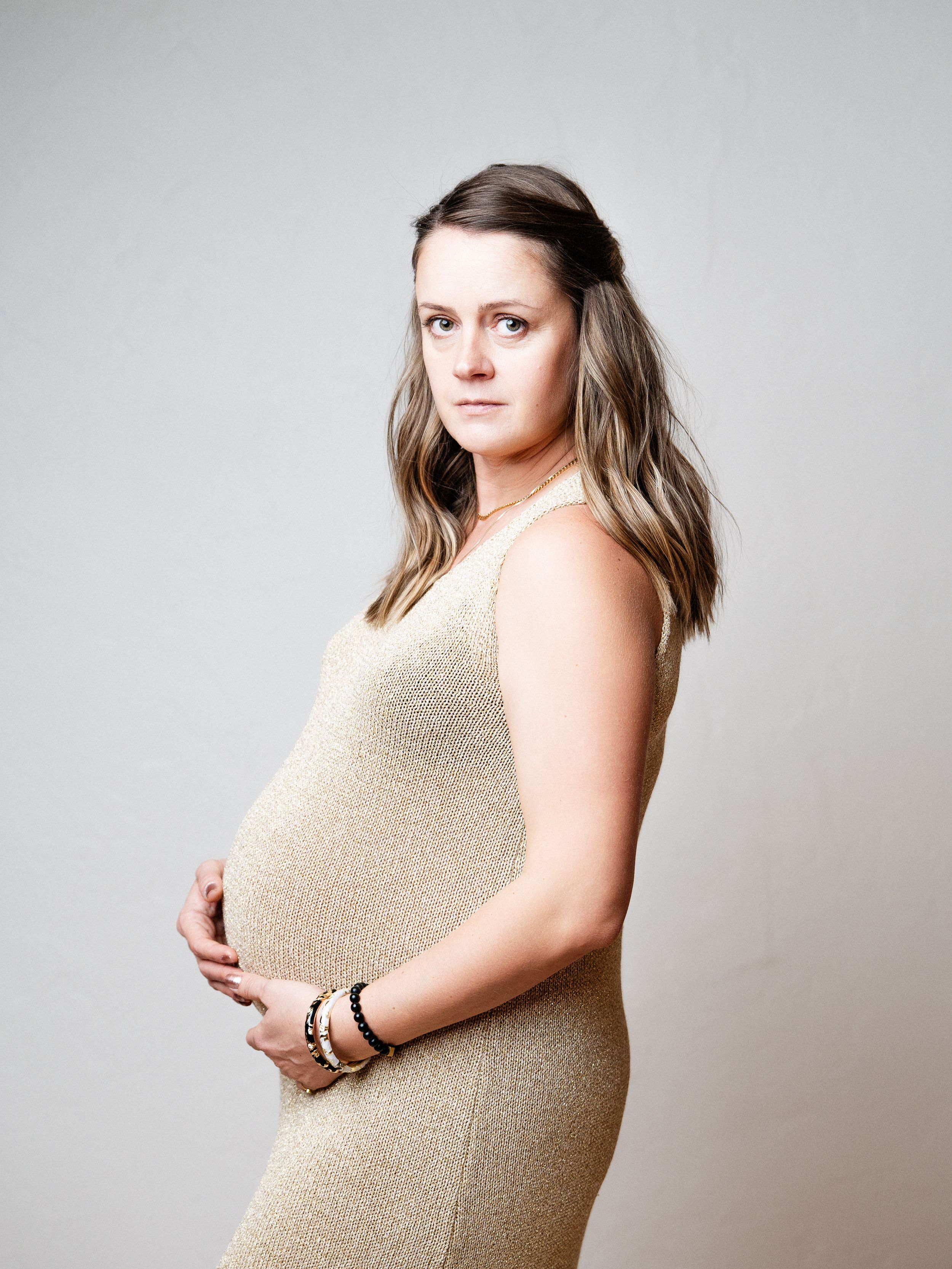 Side view maternity portrait featuring figure in beige fitted dress against neutral backdrop.