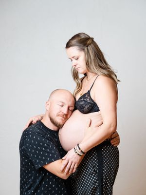 A couple shares an intimate maternity photo with expecting mother in black lace and partner embracing her belly against gray backdrop.