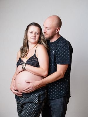 Intimate maternity photo of expectant couple embracing against gray background, with woman in lace top and man in patterned shirt.