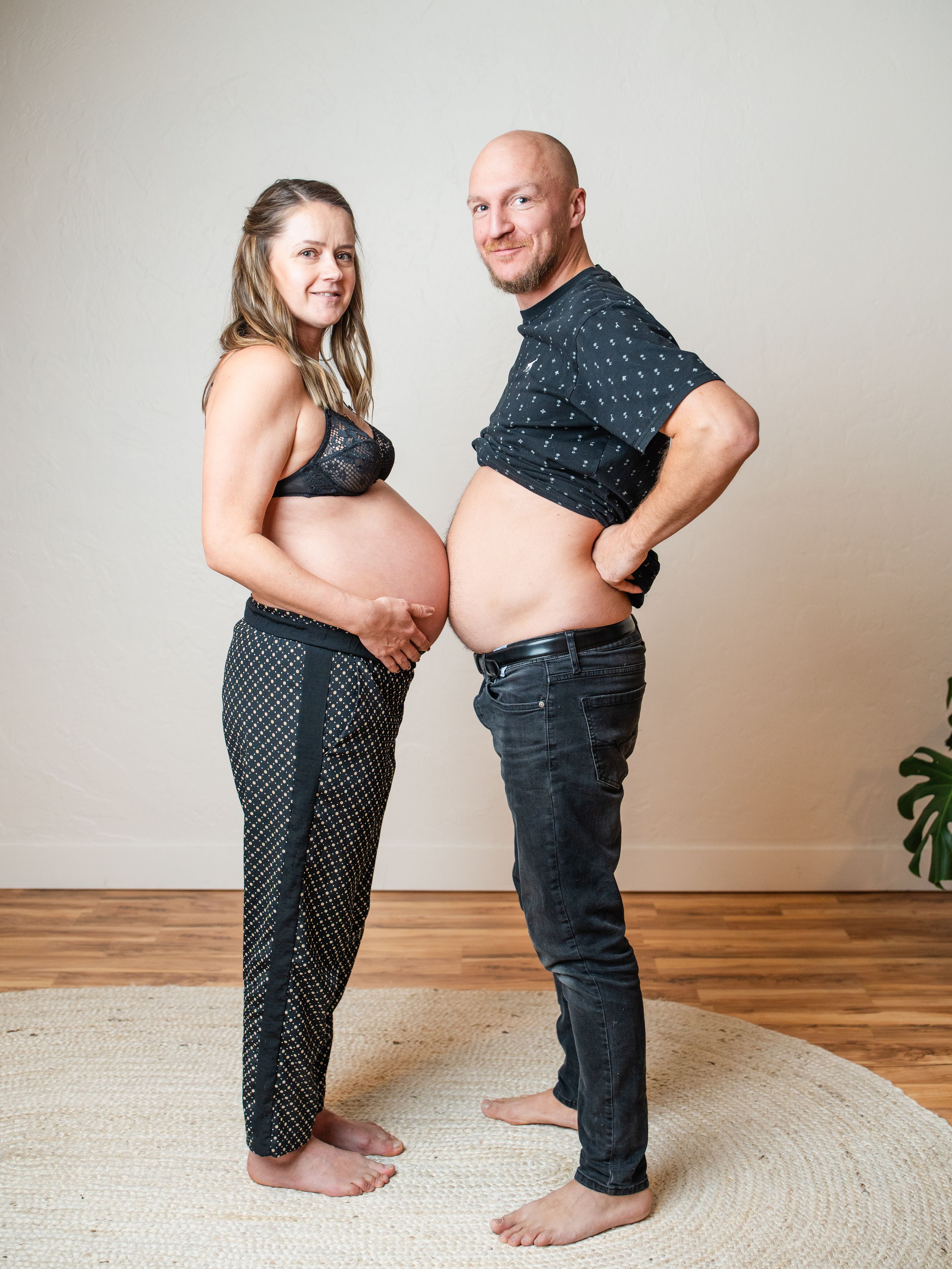Expectant couple in dark clothing pose playfully comparing their bellies in a lighthearted maternity photo.
