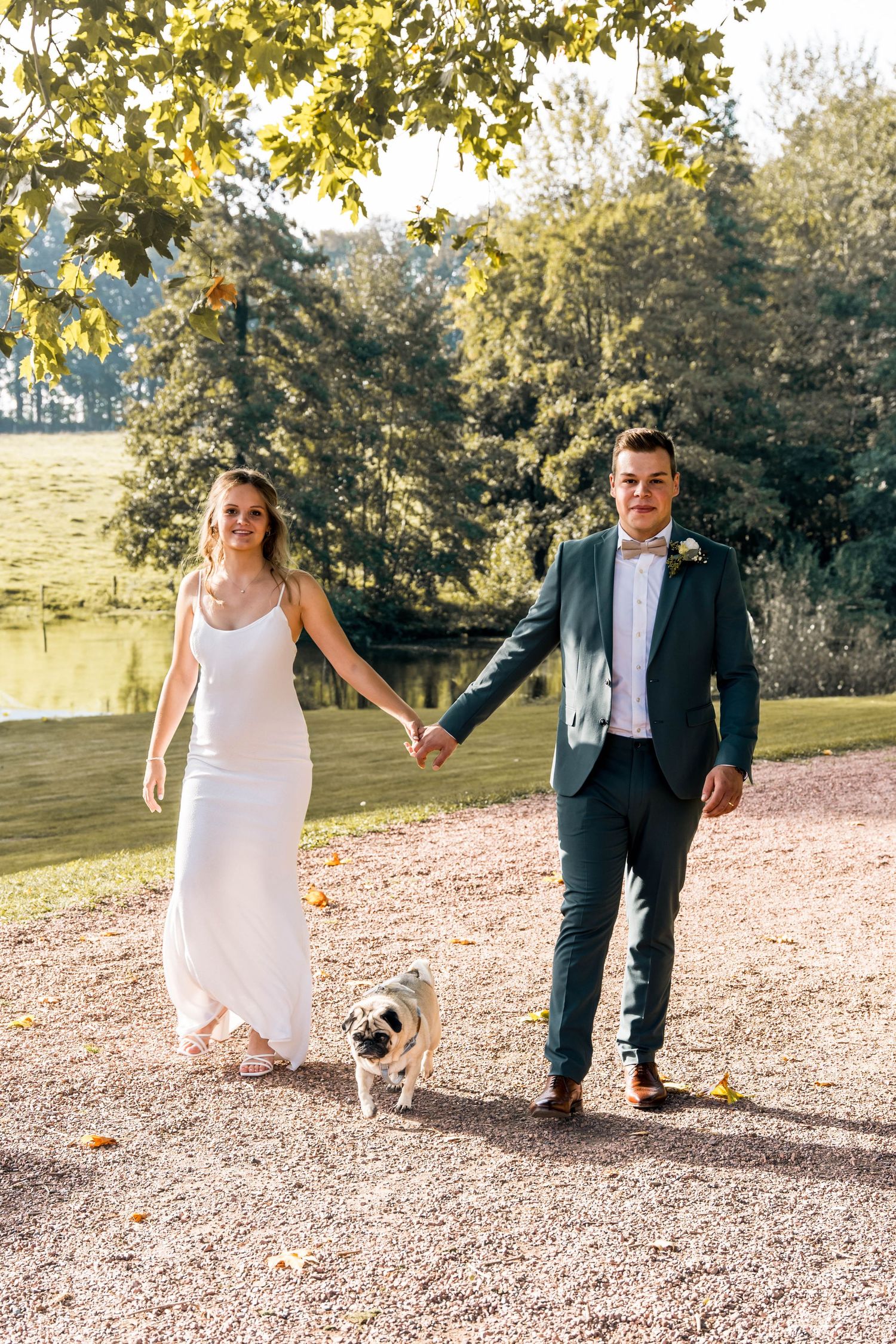 Wedding couple walking with small dog on gravel path near pond with autumn trees.