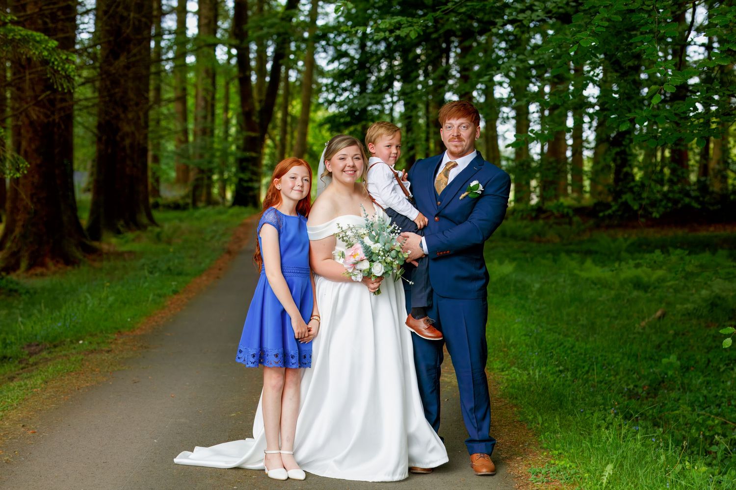 Wedding family portrait on a tree-lined path with bride, groom, and two red-haired children in blue attire.