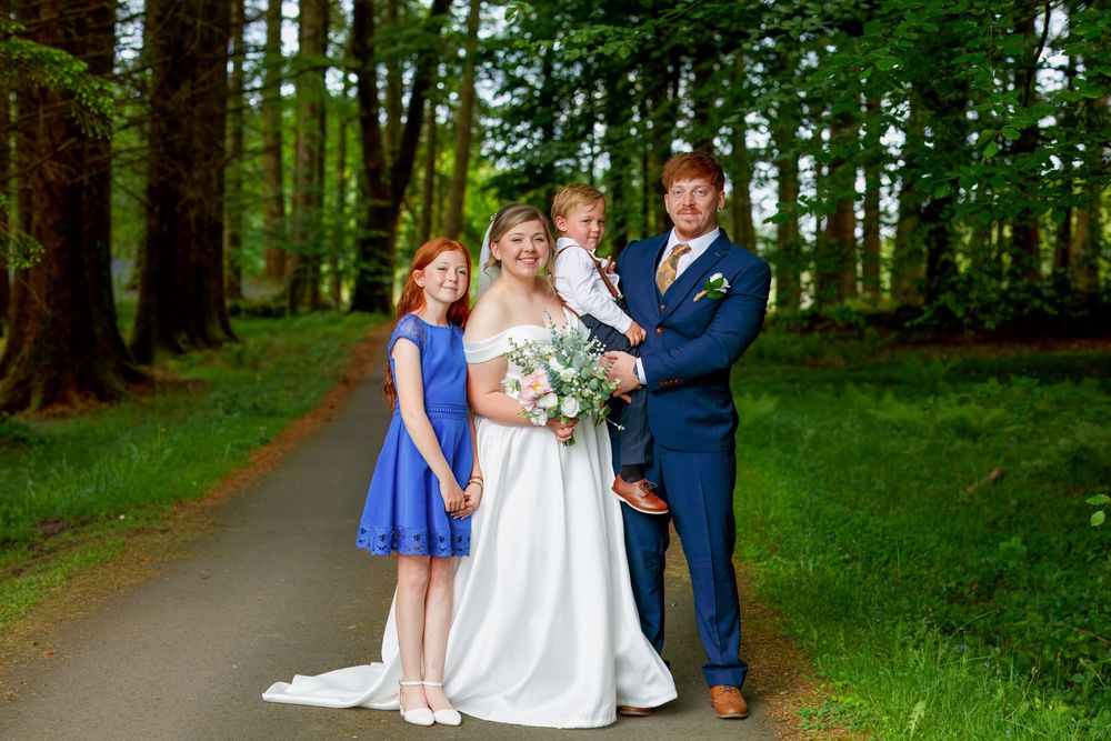 Wedding family portrait on a tree-lined path with bride, groom, and two red-haired children in blue attire.