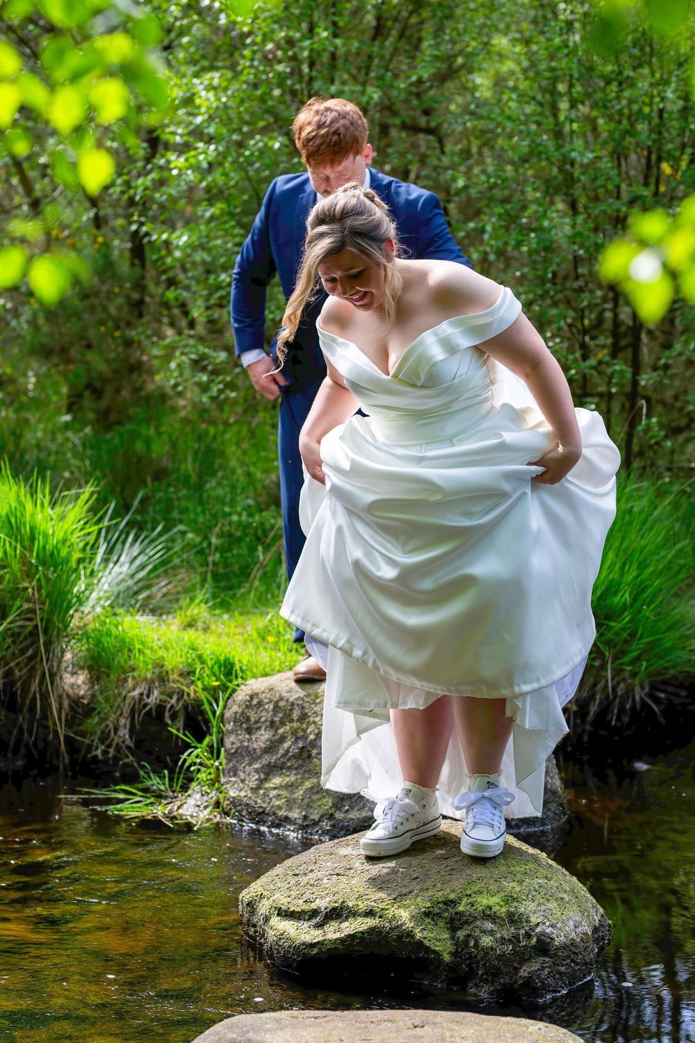 Bride in white gown and sneakers stepping on rocks over stream, groom in blue suit assisting her outdoors.