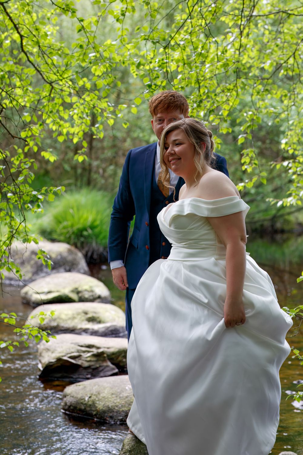 Bride in white gown and groom in blue suit walk on stepping stones by a stream in a lush green forest.
