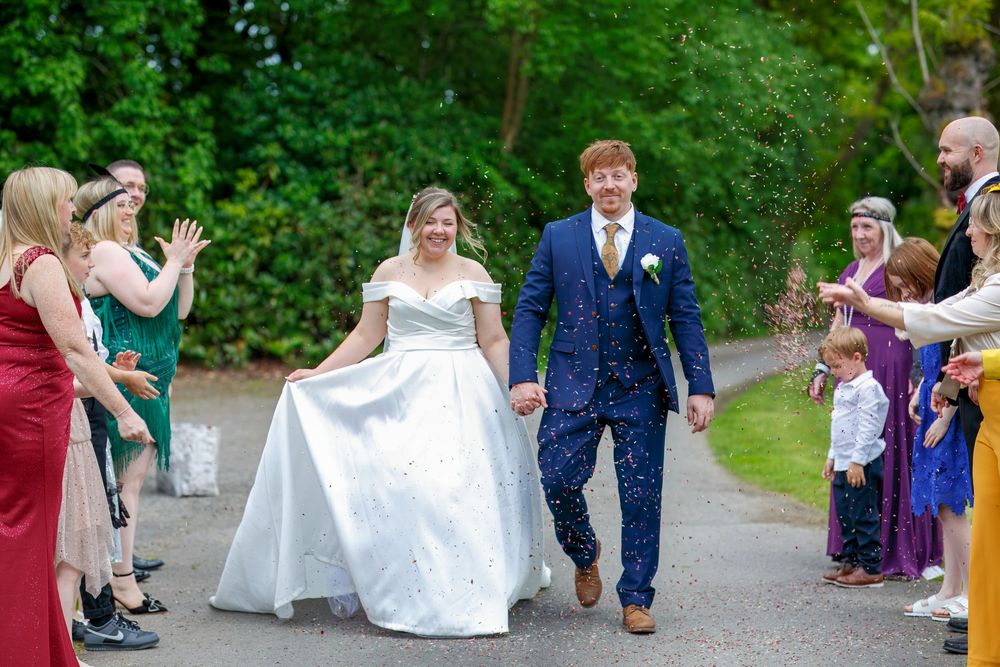 Bride and groom walk through confetti shower with smiling wedding guests lining the path outdoors.