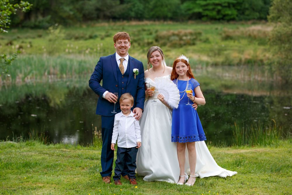 Wedding family portrait with bride, groom, young boy, and girl in blue dress beside a scenic pond.