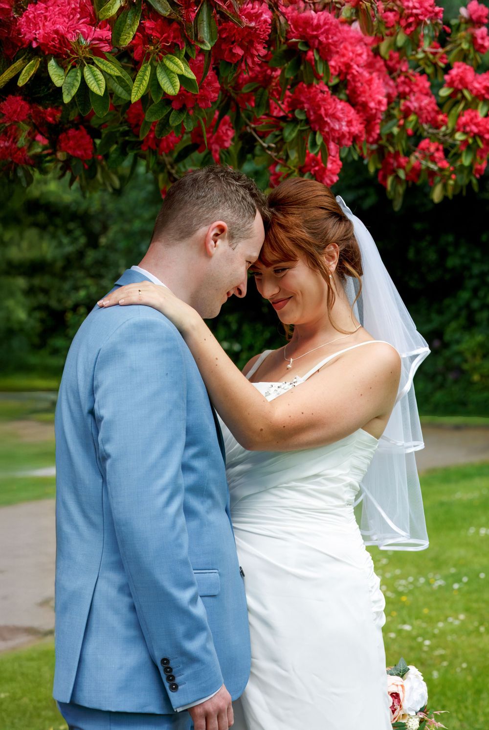 Bride in white dress and veil with groom in blue suit sharing romantic moment under blooming red flowers outdoors.