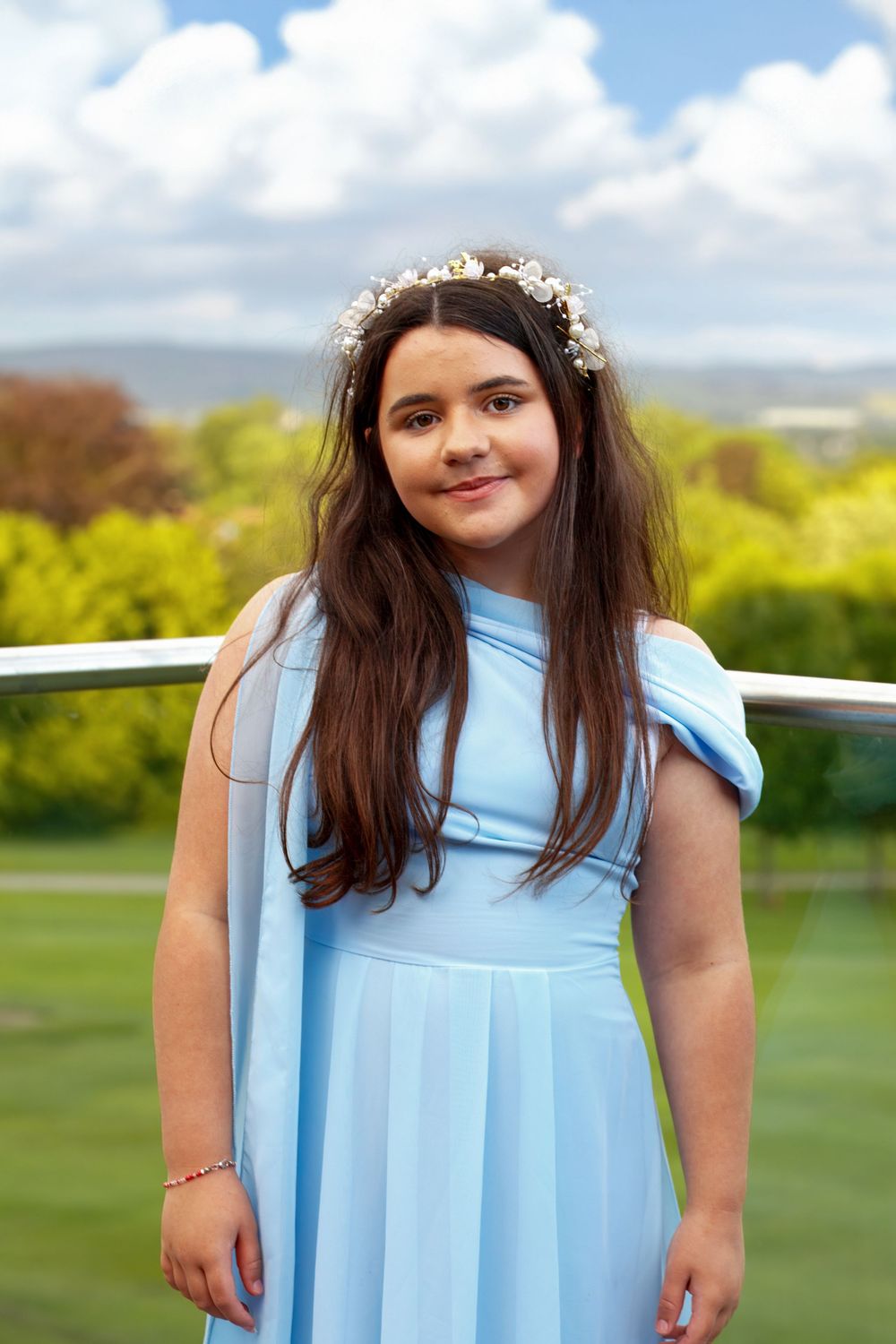 Young girl in light blue dress and floral crown smiling outdoors at a wedding celebration.
