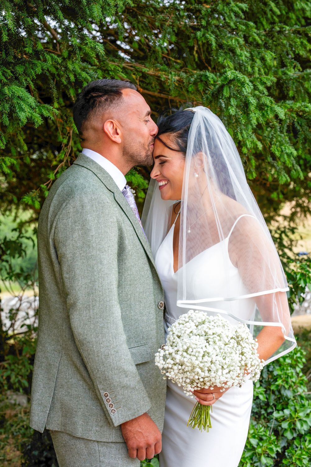 Groom in grey suit kissing bride wearing white dress and veil, holding baby's breath bouquet outdoors.
