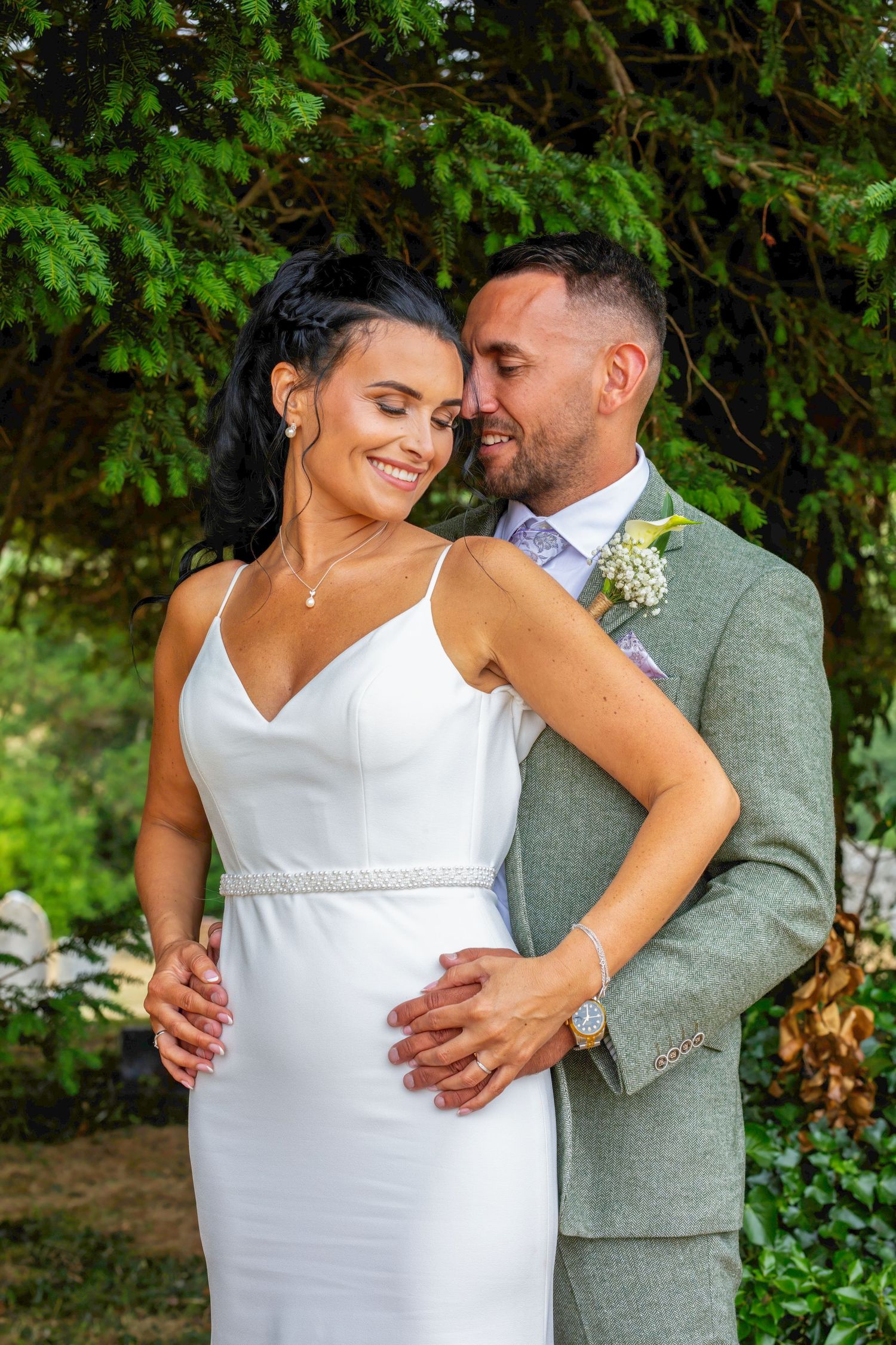 Bride in white gown and groom in sage suit embrace outdoors under lush green trees in a romantic wedding portrait.