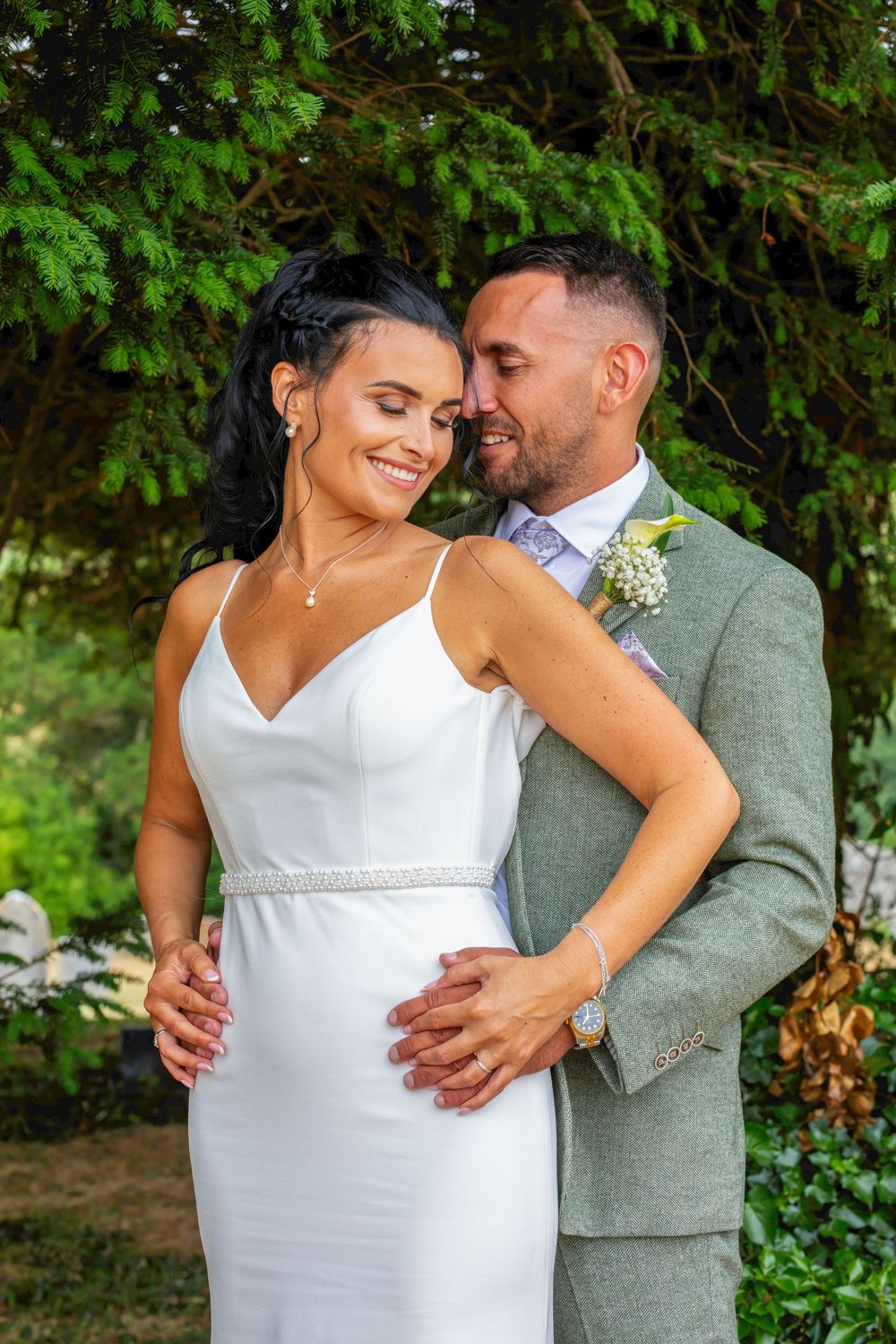 Bride in white gown and groom in sage suit embrace outdoors under lush green trees in a romantic wedding portrait.
