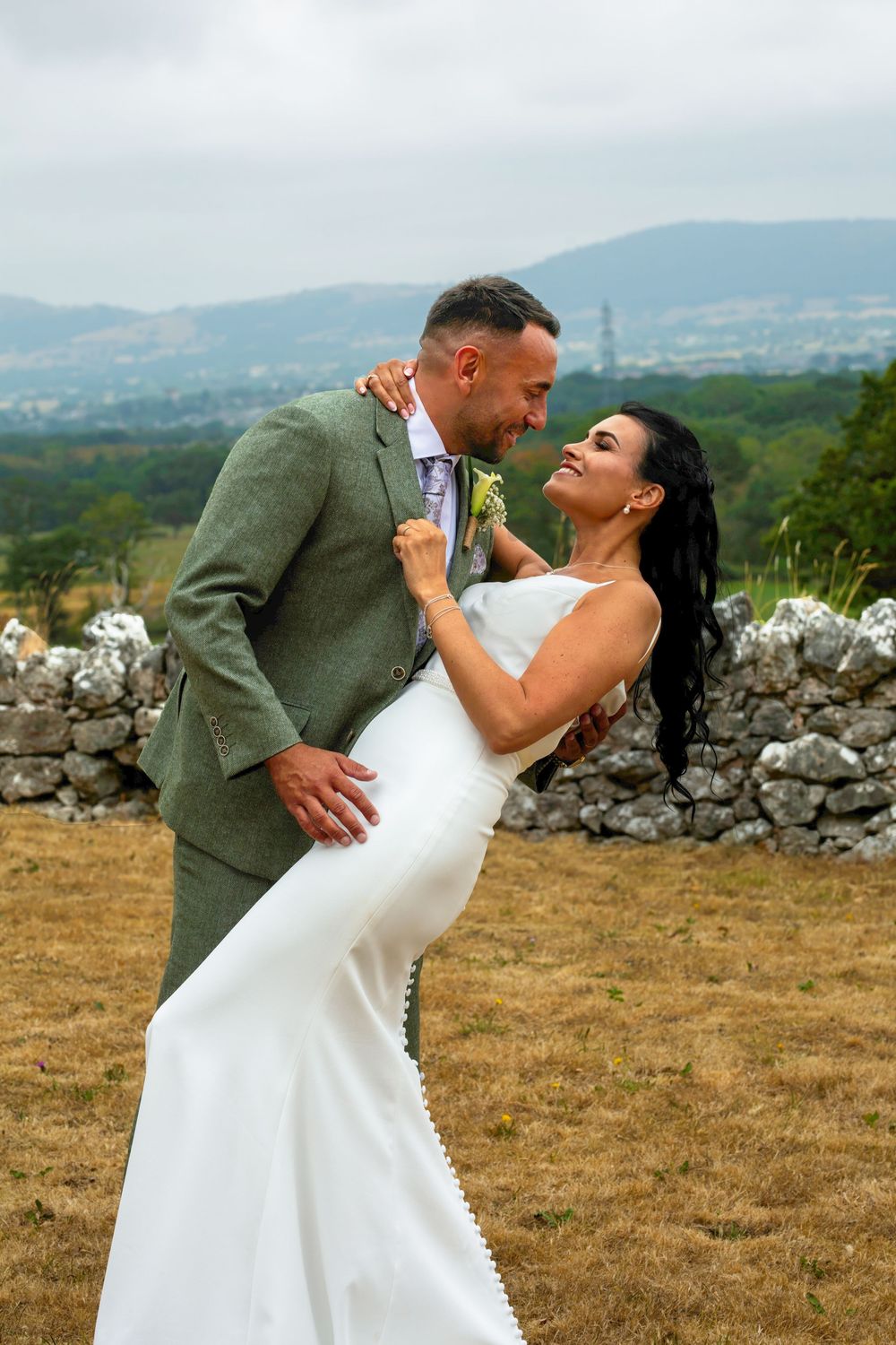 Bride and groom share a romantic dance outdoors, with scenic hills and a stone wall backdrop on their wedding day.