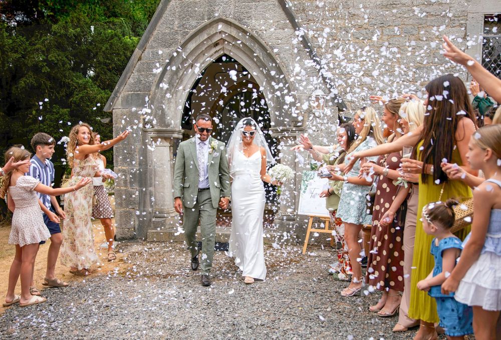 Bride and groom exit stone church amid confetti shower, guests lining path in summer wedding celebration.
