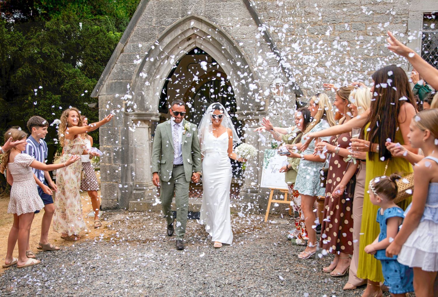 Bride and groom exit stone church amid confetti shower, guests lining path in summer wedding celebration.