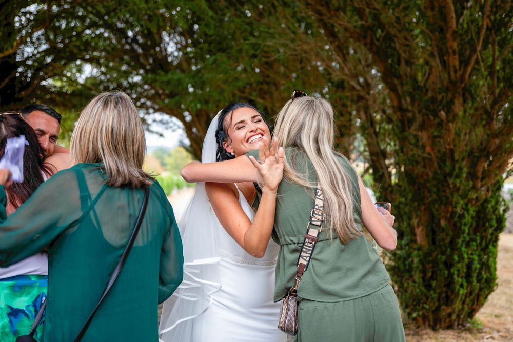 A smiling bride in white dress and veil embracing guests in green outfits outdoors at a wedding celebration.