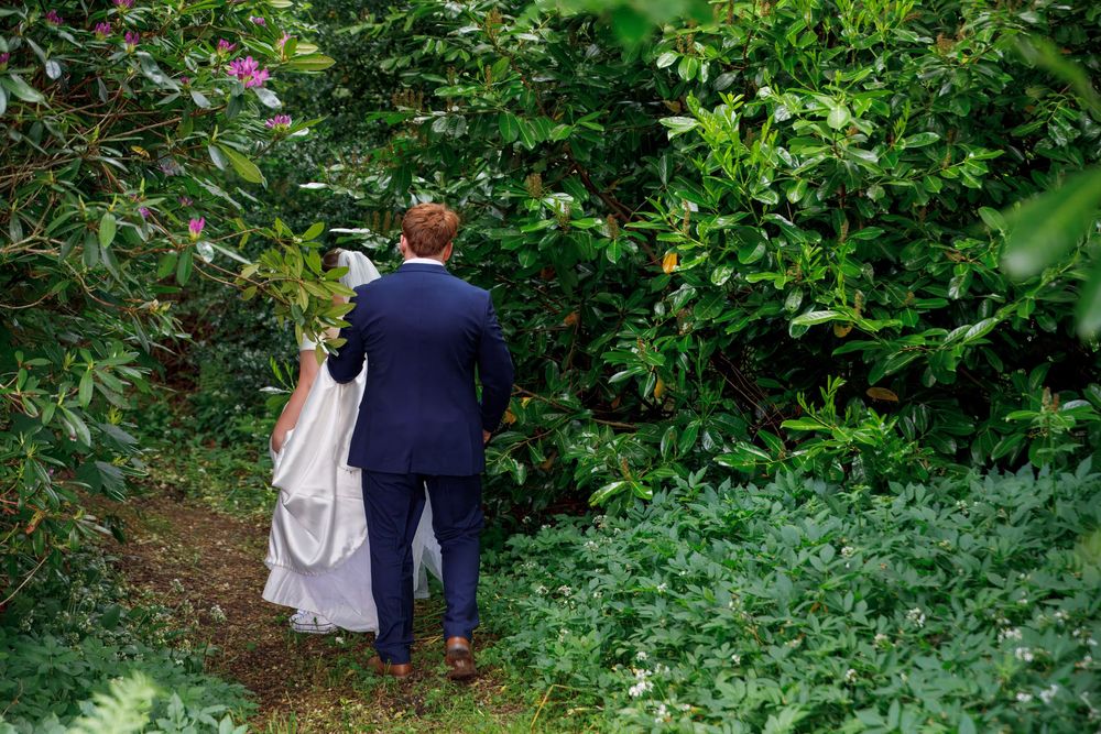 Bride and groom walk hand in hand along a lush green garden path, groom in navy suit, bride in white gown.